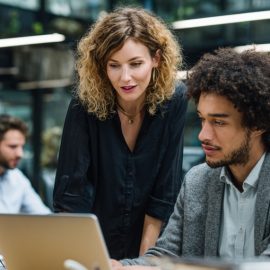 Three people in a workspace, including a manager talking to an employee at his desk, illustrates the leader-leader model