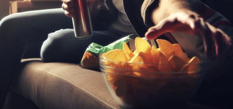 A man affected by dopamine and impulsivity as he sits on the couch and eats potato chips.