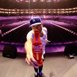 Stand up Joy Koy holding a Coca-Cola bottle on stage in an empty arena.