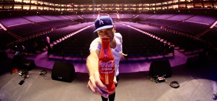 Stand up Joy Koy holding a Coca-Cola bottle on stage in an empty arena.