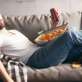 A guy lying to himself about his health as he sits on the couch with junk food and a soda can in his hand.