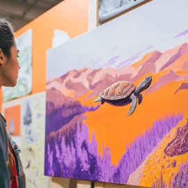 a woman looking at a painting of a turtle flying over orange mountains and purple trees illustrates the art of being wrong