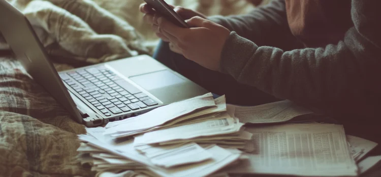 3 Reasons Why We Procrastinate (+ How to Stop) A person texting with their laptop open and papers next to them, showing one of the reasons why we procrastinate