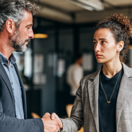 A businessman and business woman standing in an office shaking hands with unhappy looks on their faces
