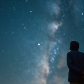 A person, seen in silhouette from behind, looking up at stars and the Milky Way in the night sky