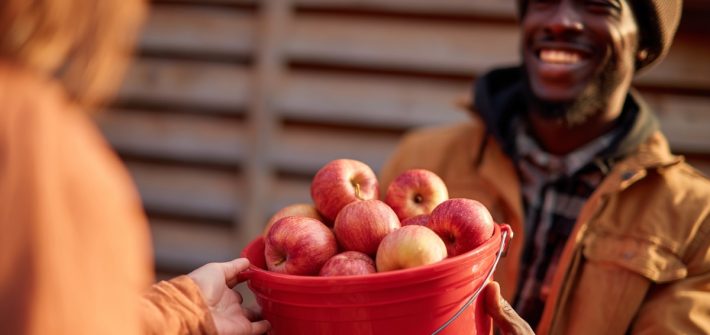 A smiling man handing a bucket of apples to a woman illustrates what a gift economy is