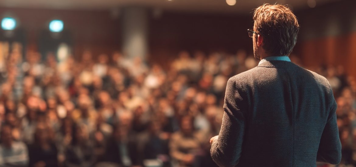 A man in a sports jacket, seen from behind, speaking to a crowd illustrates the importance of understanding your audience