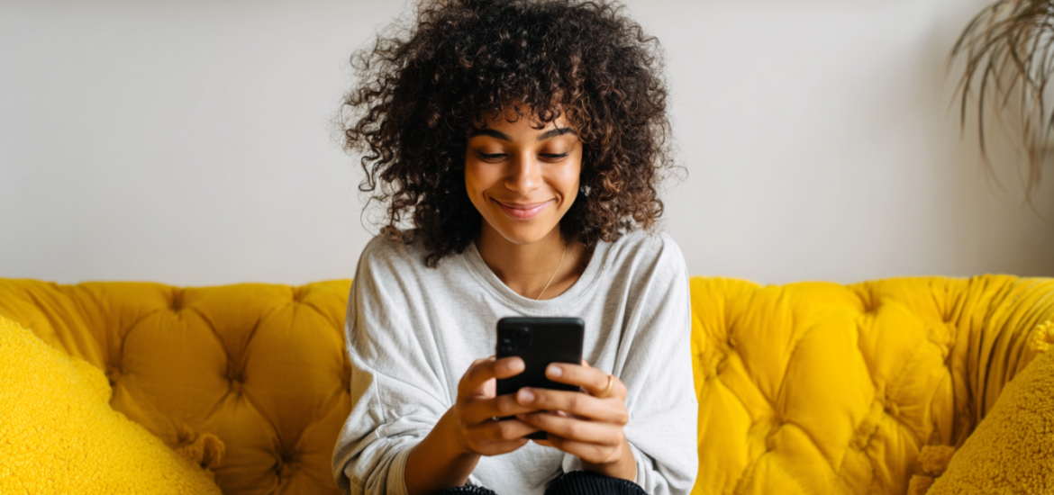 A woman smiling and using her phone while sitting on a yellow couch