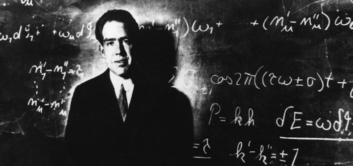 Niels Bohr standing in front of a chalkboard that has calculations written on it