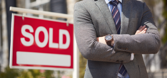 A real estate agent standing in front of a "SOLD" sign, accomplished through real estate wealth management skills