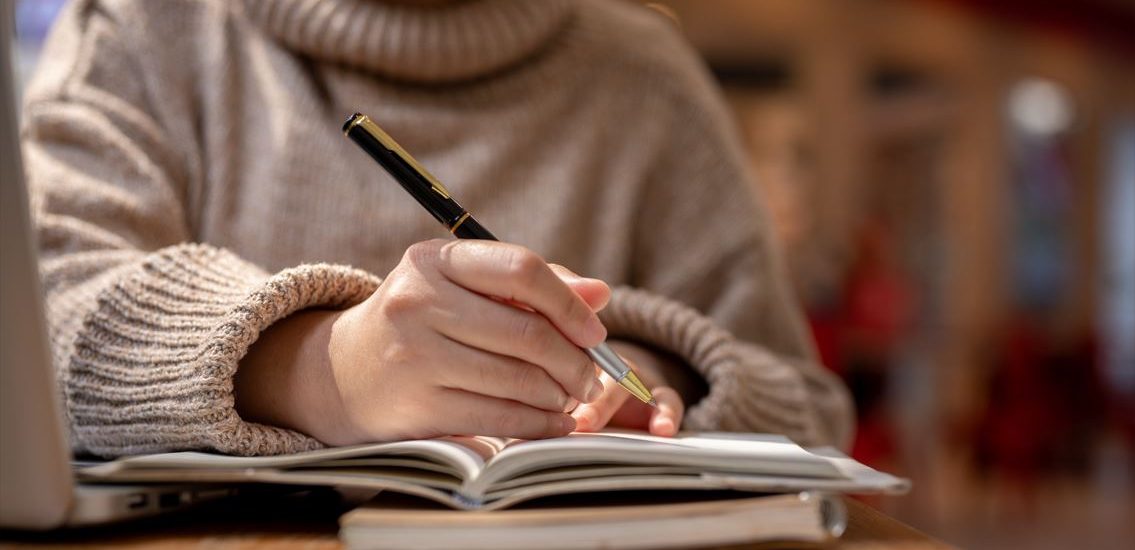 A woman in a sweater holding a pen and writing a story in a notebook