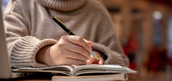 A woman in a sweater holding a pen and writing a story in a notebook