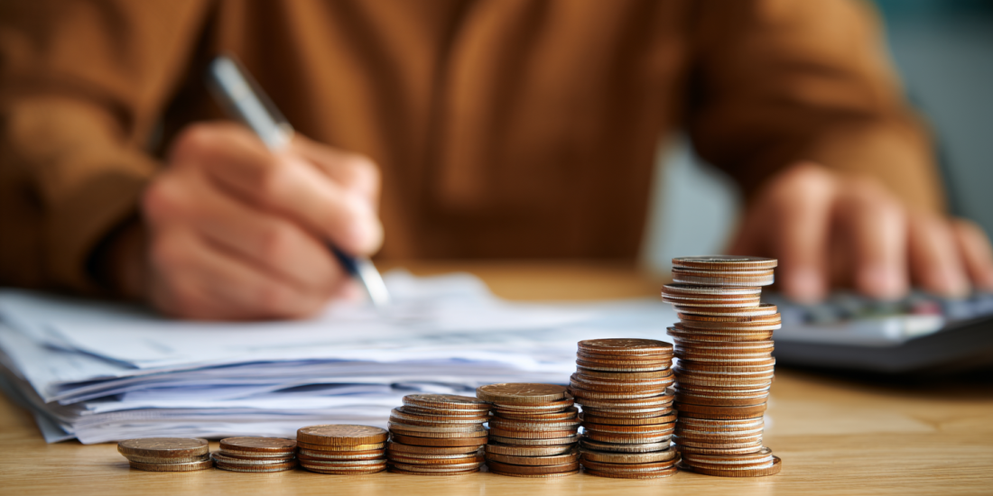 A person budgeting with a calculator behind a stack of coins